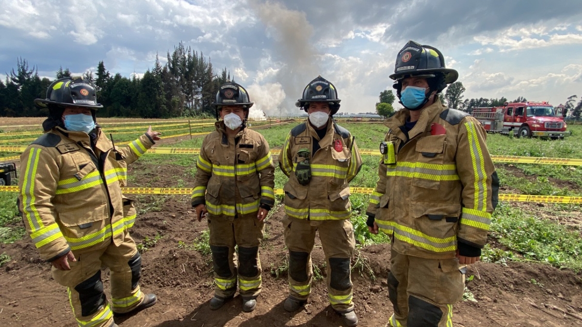 Cuatro bomberos de pie, uno de ellos señala la columna de humo con la pólvora destruida